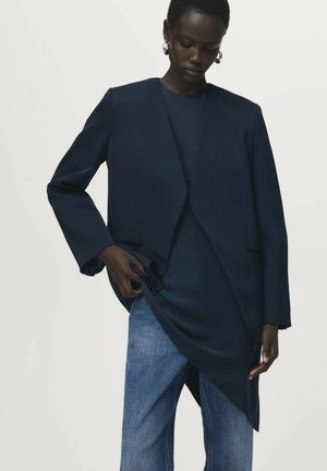 Marineblazer mit einem eleganten, modernen Schnitt; Wollmaterial, offene Vorderseite und Seitentaschen. Kombiniert mit einem dunklen Oberteil und hellen Jeans.