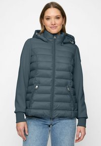 Frau in einer türkisfarbenen Steppjacke mit Reißverschluss und Taschen, Kapuze mit Kordelzügen und blauen Jeans, steht vor einem weißen Hintergrund.
