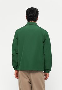 Chaqueta verde con cuello, puños elásticos y corte recto. Textura de tela suave, diseño minimalista sin patrones ni acentos visibles.