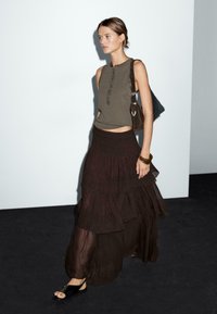 Olivegrünes, ärmelloses Oberteil mit Knopfakzenten, brauer Maxi-Rock mit Lagen und schwarze Sandalen. Das Model trägt eine dunkle Tasche. Einfaches, fließendes Design.