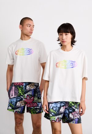 Bele majice s večbarvnim logotipom "adidas", v kombinaciji z vzorčastimi kratkimi hlačami, ki vsebujejo živahne grafike in logotipe v zeleni, vijolični in modri barvi.
