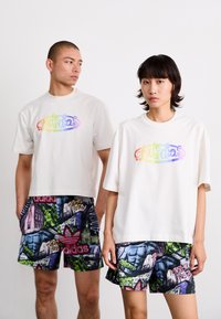 Hvide t-shirts med multicolore "adidas" logo, parret med mønstrede shorts med farverige grafikker og logoer i grøn, lilla og blå.