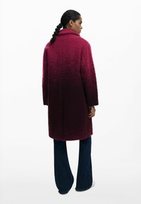 Bordeauxfarbener, strukturierten Mantel mit Kragen und langen Ärmeln, aus einem weichen, flauschigen Stoff. Kombiniert mit weit geschnittenen, dunklen Jeans.