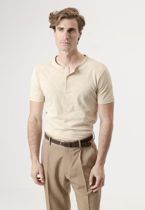 Joven con cabello castaño claro que lleva una camiseta henley beige de manga corta y pantalones marrones con un cinturón oscuro, de pie frente a un fondo liso.