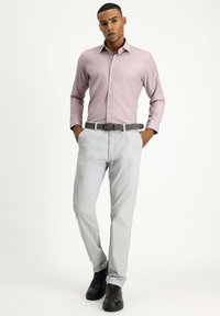 Camicia button-up rosa chiaro con maniche lunghe, abbinata a pantaloni grigi chiari e una cintura grigia a fantasia. Le scarpe nere completano l'outfit.