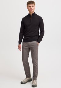 Svart, långärmad, zip-up tröja med bruna detaljer, kombinerad med ljusgrå byxor och grå vandringsskor, stående mot en neutral bakgrund.