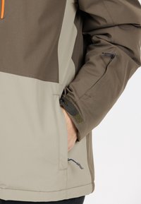 Brun och beige softshell-jacka med dragkedjefickor och kardborrefästen. Har en orange accent och en texturerad tygdesign.