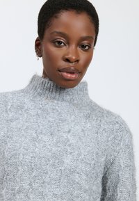 Kvinde med kort krøllet hår iført en lysegrå højhalset kabelstrikket sweater og små øreringe, som ser direkte ind i kameraet.