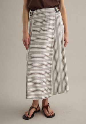 Donna che indossa una gonna a portafoglio alla caviglia con righe marroni orizzontali e verticali, abbinata a sandali marroni e un top scuro.