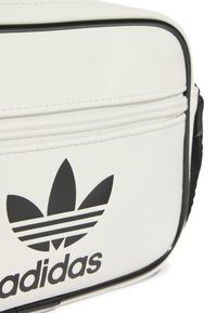 Bílá syntetická taška s černým logem Adidas se třemi pruhy. Obsahuje zipové uzavření a kontrastní černé detaily. Texturovaný povrch.