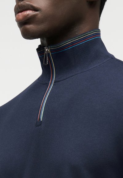 Μπλε ναυτικό πουλόβερ με ψηλό γιακά, με μισό φερμουάρ και πολύχρωμες ριγέ λεπτομέρειες κατά μήκος του γιακά. Μαλακό, υφασμάτινο με υφή.