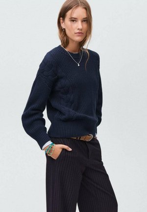 Maglione di colore blu marina a maglia con design a trecce testurizzate, polsini e orlo a costine. Abbinato a pantaloni a righe e braccialetti sovrapposti.