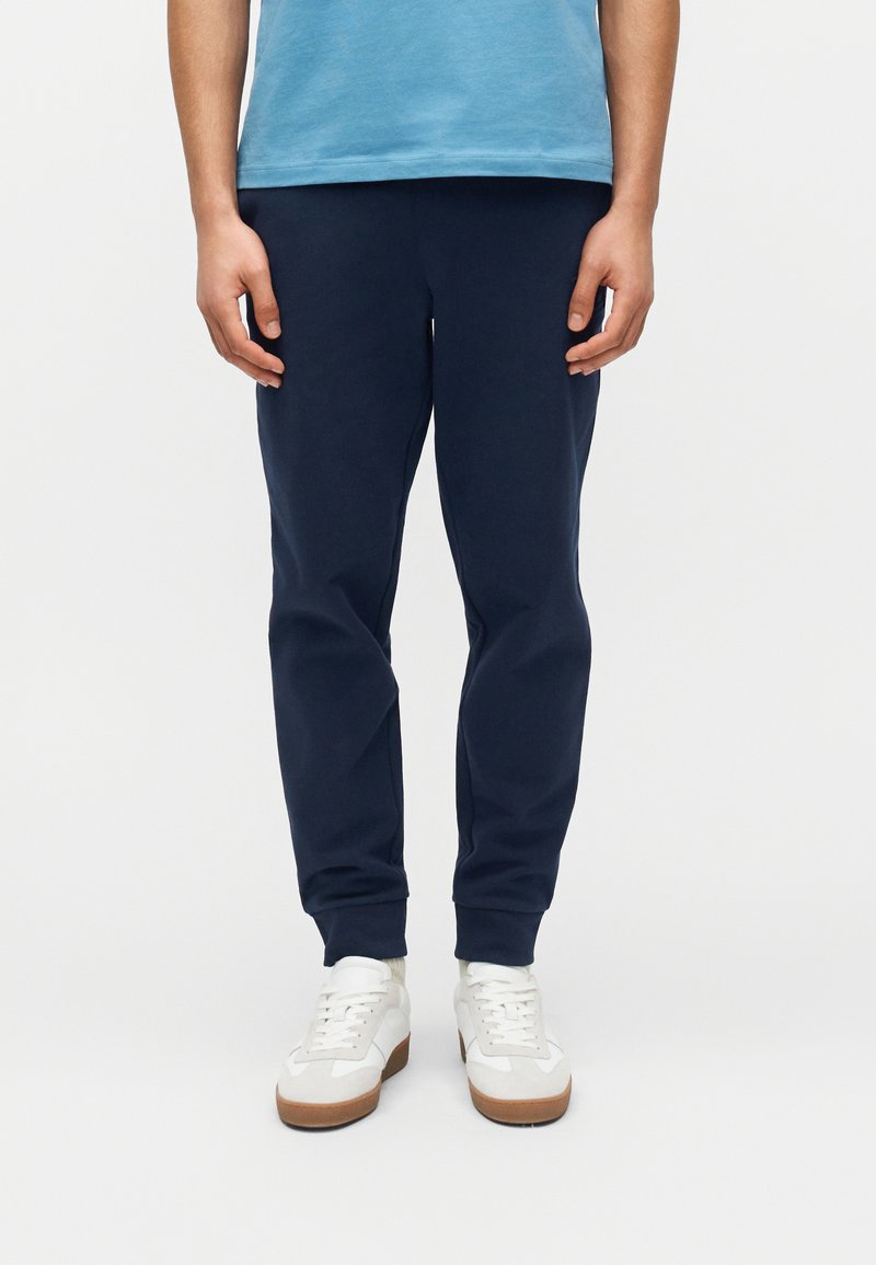 Άτομο που φοράει μπλε σκούρο παντελόνι τύπου jogger, λευκά αθλητικά παπούτσια με καφέ σόλες και γαλάζιο πουκάμισο, στέκεται μπροστά σε απλό λευκό φόντο.