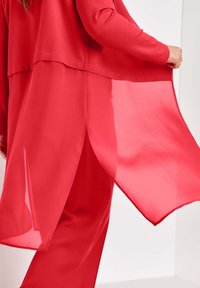 Persona con una camiseta de manga larga roja y pantalones, sosteniendo el tejido ligero y fluido del panel lateral de la camiseta.