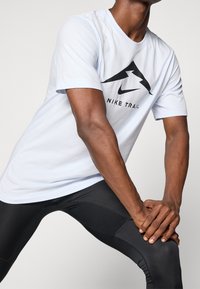 Světle modré tričko s krátkým rukávem s černým logem hory a nápisem "NIKE TRAIL", doplněné černými přiléhavými legínami. Hladká textura látky.