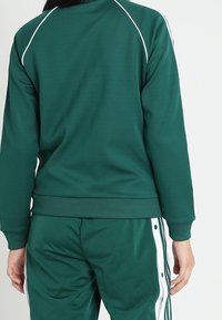 Zelená lehká atletická bunda se světlými bílými linkami na ramenou. Má hladkou texturu a přiléhavý střih, doplněná o zelené kalhoty stejného odstínu.