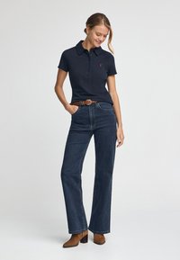 Navyblaues Polohemd mit Kragen, vier Knöpfen und kleinem Logo. Kombiniert mit hochgeschnittenen, weit geschnittenen dunkelblauen Jeans und braunen Stiefeletten mit Absatz.