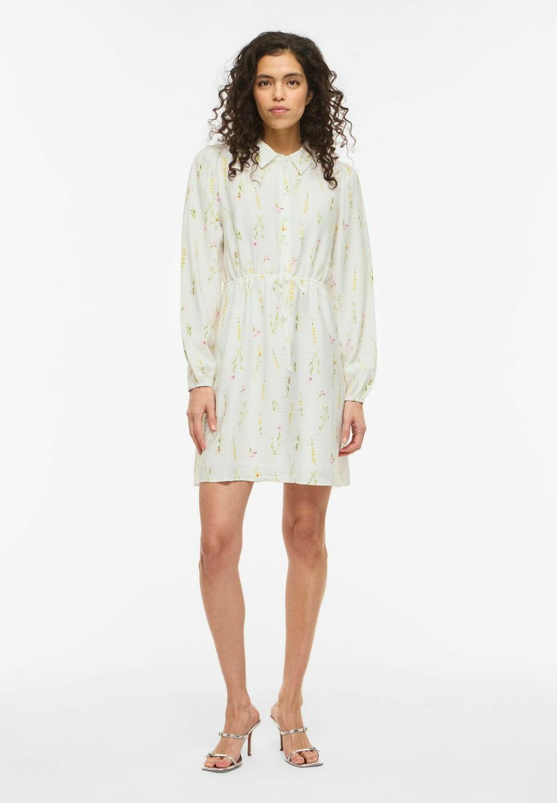 Kiharaliste juustega naine, kes kannab valget lillemustriga pikkade varrukatega kleiti, mis on vöökohast kitsendatud, ja hõbedaseid kõrgete kontsadega sandaale, seistes valge tausta ees.