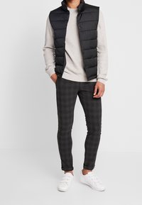 Black puffer vest over a light gray textured sweater, paired with dark gray check-patterned trousers and white sneakers.
