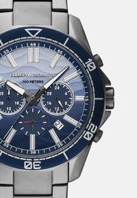 Srebrny zegarek z nierdzewnej stali z niebieską obręczą i teksturowaną niebieską tarczą, z trzema subtarczami i wyświetlaczem daty na pozycji 6.