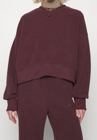 Mörkröd fleece-tröja med kortad design, långa ärmar, ribbade muddkanter och en svart Nike-logotyp på bröstet. Slät textur.