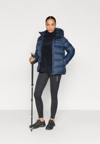 Marineblå dunjakke med hætte, båret over en mørk sweater og stramme sorte leggings. Sorte vandresko og en vandrestav fuldender outfittet.