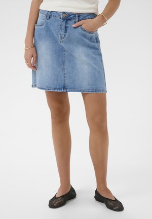 Kvinde iført lys blå denims nederdel, hvid top, sorte flettede flade sko og guldbøjler med hånden i lommen mod en ensfarvet baggrund.