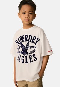 Baltas kokvilnas T-krekls ar tumši zilu grafikas attēlu, kurā redzams ērglis un uzraksts "SUPERDRY EAGLES". Īsas piedurknes, brīvs piegriezums, ar sarkaniem akcentiem.