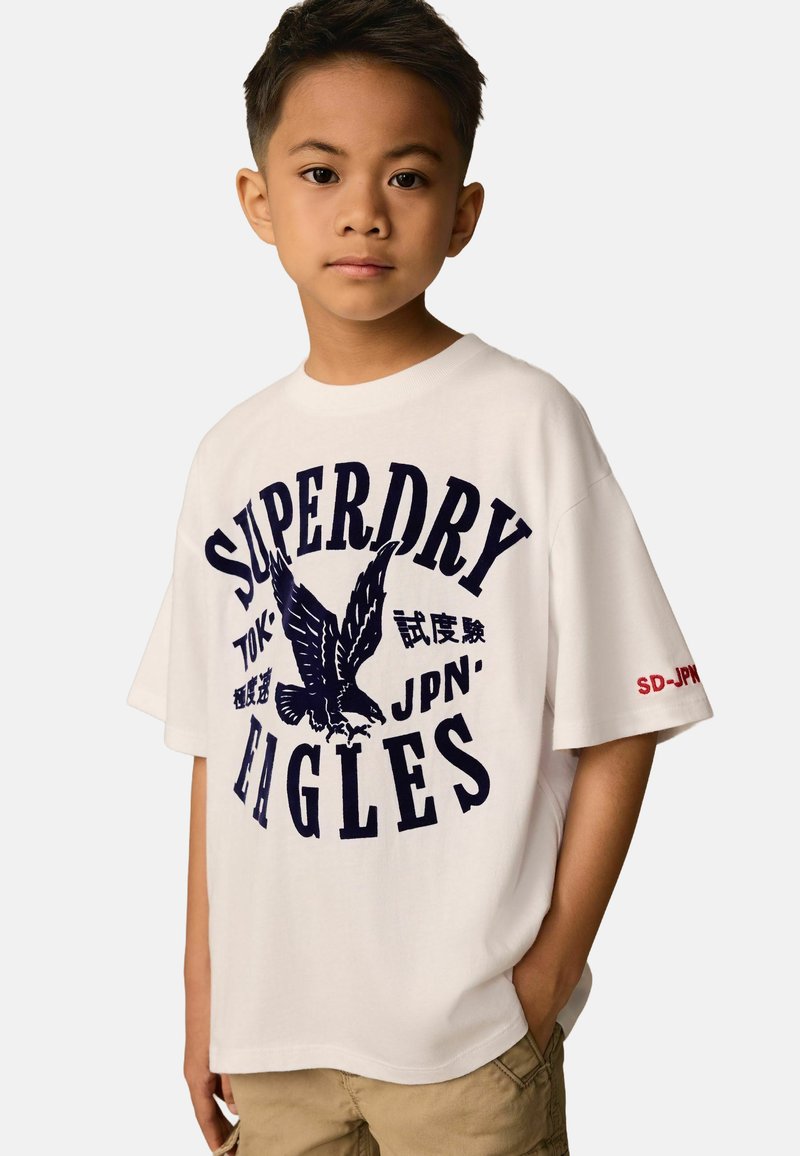 Baltas kokvilnas T-krekls ar tumši zilu grafikas attēlu, kurā redzams ērglis un uzraksts "SUPERDRY EAGLES". Īsas piedurknes, brīvs piegriezums, ar sarkaniem akcentiem.