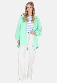 Mentazöld vízálló dzseki, bő szabású kapucnival, kék-fehér csíkos ing és világos kék farmer széles szárú nadrág fölött viselve.