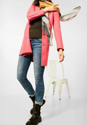 Femme portant un manteau rose, une écharpe colorée, un jean bleu et des bottes noires, debout près d'une chaise en métal blanche sur un fond uni.