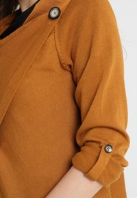 Mosterdgele gebreide cardigan met een wikkelontwerp, voorzien van een zwarte knoopdetail en opgerolde mouwen. Zachte textuur met een gladde afwerking.