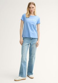 Camiseta de manga corta azul claro con el texto blanco "EL MÁS FELIZ JUNTO AL MAR", combinada con unos jeans acampanados azul claro y zapatillas blancas.