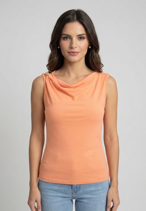 Donna con capelli scuri che indossa una maglia pesca senza maniche con scollo arricciato e fibbia dorata, abbinata a jeans azzurri, in piedi davanti a uno sfondo semplice.