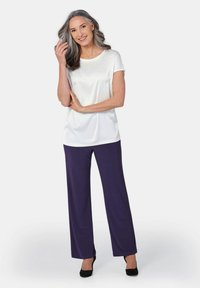 Weiße, seidene Kurzarmbluse mit dezenten Verzierungen, kombiniert mit lockeren lila Hosen und schwarzen Absätzen. Das Model hat lange, wellige graue Haare.