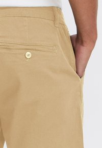 Chino bej din bumbac neted; design cu pantalon drept; buzunar spate cu închidere prin buton; buzunare laterale pentru mâini.