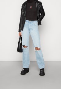 Person som bär en svart läderjacka, svart turtleneck med logotyp, ljusblå slitna jeans, svarta plattformsstövlar och håller en svart handväska.