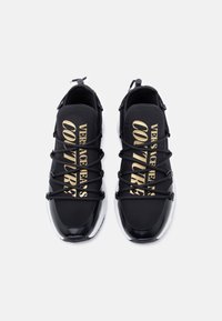 Μαύρα αθλητικά παπούτσια με ελαστικά κορδόνια, με χρυσή επωνυμία «VERSACE JEANS COUTURE» στο πάνω μέρος. Υφή στη μαύρη και λευκή σόλα.