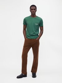 Uomo con maglietta verde a maniche corte con taschino sul petto e pantaloni marroni, in piedi con le mani nelle tasche davanti a uno sfondo semplice.