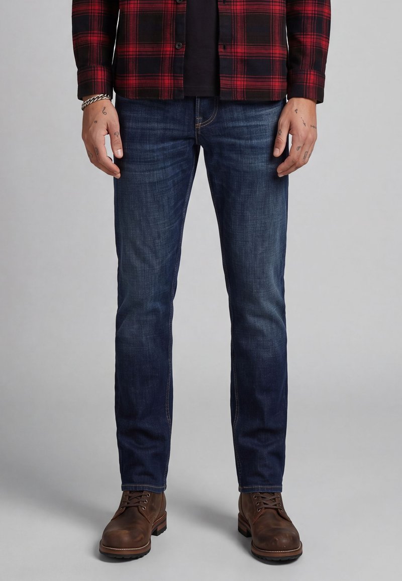 Man draagt donkerblauwe slim-fit jeans, bruine leren laarzen en een rood-zwart geruit overhemd, staand tegen een neutrale achtergrond.