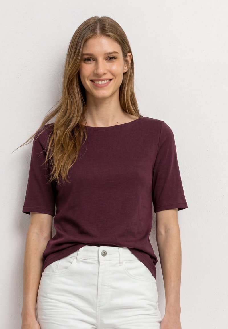 Jeune femme aux longs cheveux bruns portant un t-shirt bordeaux à manches courtes et un pantalon blanc, souriant devant un fond clair uni.