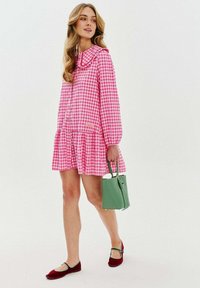 Růžové kostkované šaty s volánkovým límečkem a dlouhými rukávy, s rozšířenou sukní. Outfit doplňuje zelená kabelka a červené baleríny.