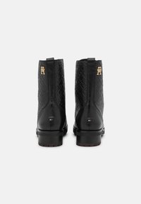 Botas de tobillo de cuero negro con un patrón texturizado, acentos del logotipo en dorado y un tacón cuadrado. La vista trasera muestra detalles y construcción.