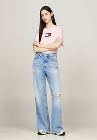 Camiseta rosa claro de manga corta con un logotipo rectangular, combinada con unos vaqueros de pierna ancha desgastados de tiro alto en azul claro y zapatillas deportivas.