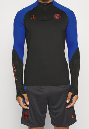 Chemise noire à manches longues avec épaules bleues, dotée d'un zip quart, d'accents orange et de manches noires texturées. Associée à un short gris.