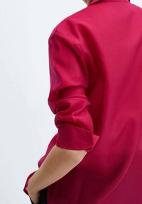 Personne portant une chemise rose vif à manches longues avec les manches retroussées, main posée sur la hanche, sur un fond clair uni.