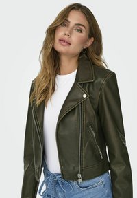 Giacca da biker in ecopelle verde oliva con accessori in argento, tasche con zip e collo a pistagna, abbinata a una maglietta bianca e jeans blu.