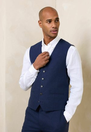 Man adjusting collar wearing navy blue vest and white button-up shirt, standing against a beige wall with one hand in pocket.