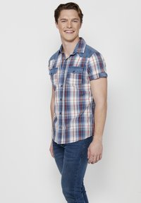 Camicia a maniche corte a quadri nei colori blu, rosso e bianco, con accenti sulle spalle simili al denim e due tasche sul petto. Indossata con jeans blu.