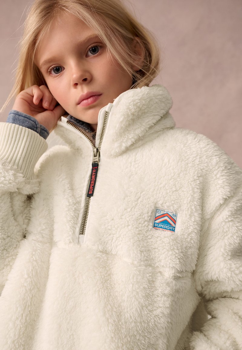 Mladé dievča so svetlými vlasmi nosí bielu plyšovú Superdry bundu na zips s vysokým golierom na jednoliatom pozadí.